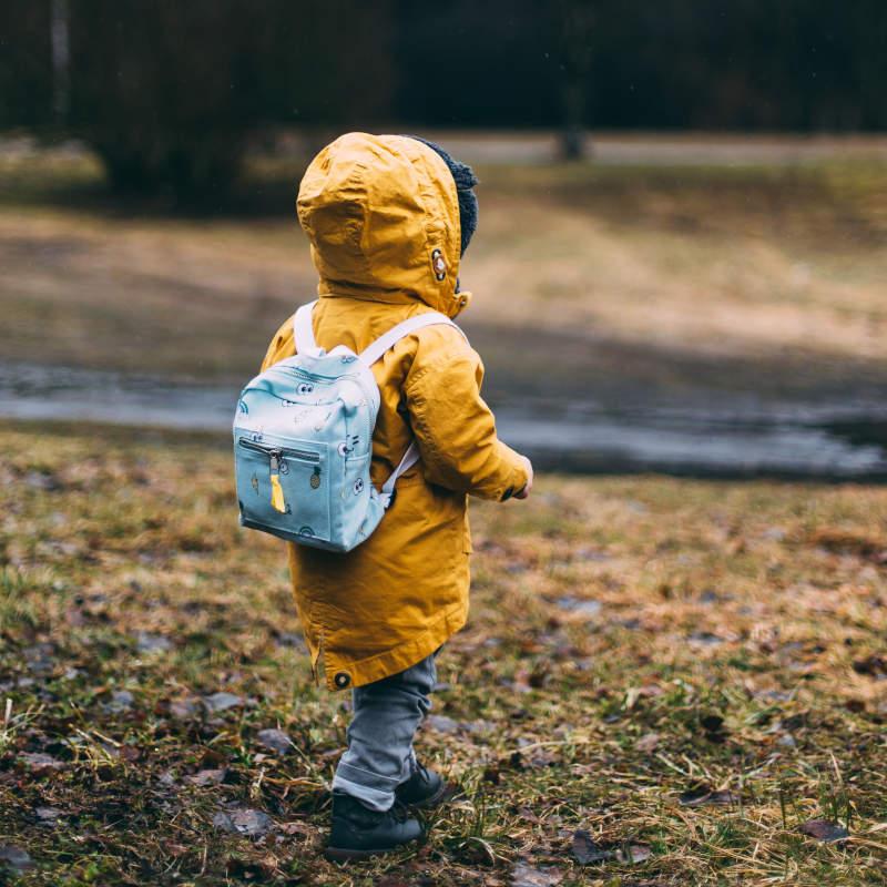Child in a yellow parka style jacket, carrying a blue backpack, walking outside.